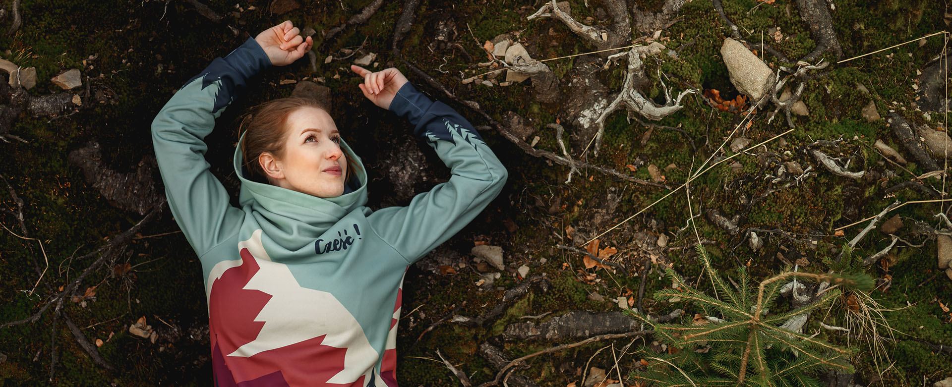 Frau liegt auf moosigem Waldboden in einem Hoodie mit Bergmotiv und weiter Kapuze
