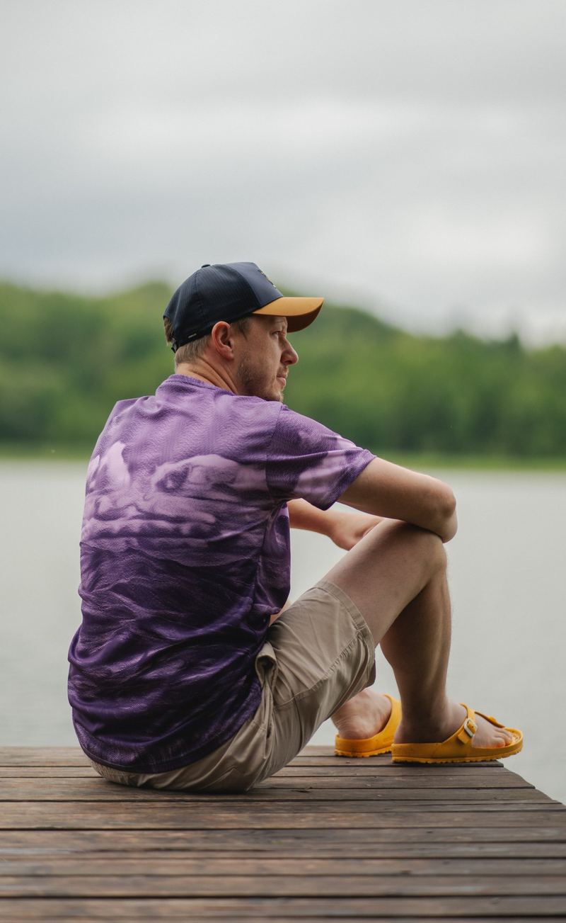 Mann auf einem Steg am See in einem lila gemusterten Shirt